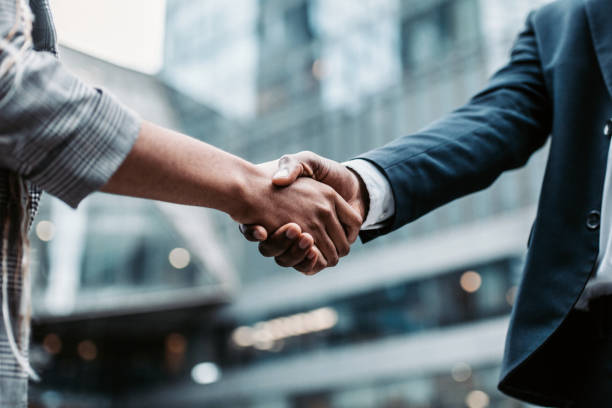 Meeting success. Two business persons shaking hands standing outside Dos personas vestidas con ropa de trabajo se dan la mano al aire libre, con edificios de oficinas al fondo.