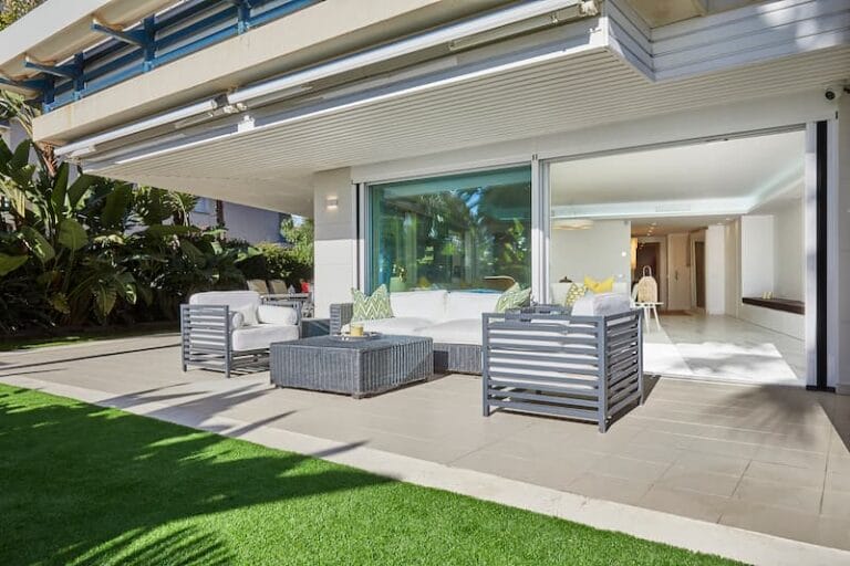 Un patio moderno con muebles de exterior grises, una mesa baja y un conjunto de sofás blancos sobre un suelo de baldosas, junto a una gran puerta corredera de cristal y rodeado de una exuberante vegetación. Un patio moderno con muebles de exterior grises, una mesa baja y un conjunto de sofás blancos sobre un suelo de baldosas, junto a una gran puerta corredera de cristal y rodeado de una exuberante vegetación.