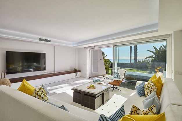 Salón moderno con un sofá modular blanco, cojines de colores, un televisor grande y puertas correderas de cristal que dan a un patio con vistas a las palmeras y al mar. Salón moderno con un sofá modular blanco, cojines de colores, un televisor grande y puertas correderas de cristal que dan a un patio con vistas a las palmeras y al mar.