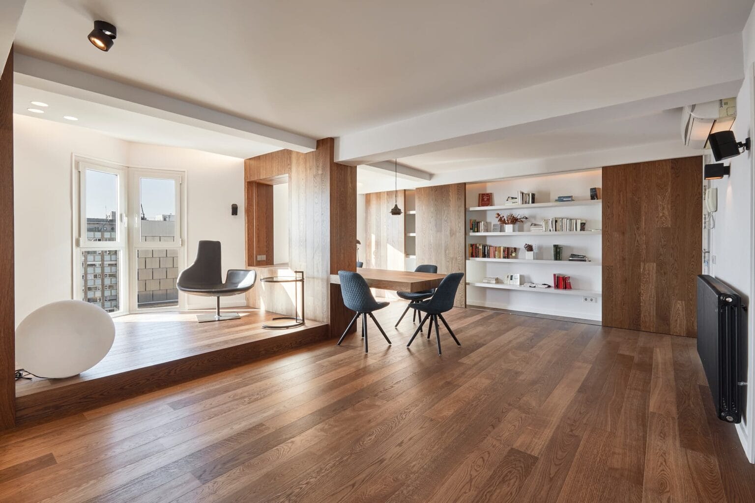 Sala de estar y comedor de estilo moderno con suelos de madera, estanterías empotradas, una mesa redonda con cuatro sillas, un sillón decorativo junto a la ventana y amplios ventanales que dejan entrar luz natural. Sala de estar y comedor de estilo moderno con suelos de madera, estanterías empotradas, una mesa redonda con cuatro sillas, un sillón decorativo junto a la ventana y amplios ventanales que dejan entrar luz natural.