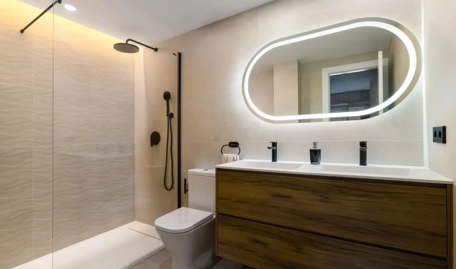 Cuarto de baño moderno con ducha a ras de suelo, cabezal de ducha tipo lluvia suspendido, inodoro, mueble de baño de madera, lavabos dobles y un espejo ovalado retroiluminado. Cuarto de baño moderno con ducha a ras de suelo, cabezal de ducha tipo lluvia suspendido, inodoro, mueble de baño de madera, lavabos dobles y un espejo ovalado retroiluminado.